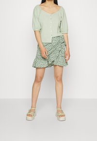 Haut vert clair à manches bouffantes avec fermeture à boutons, assorti à une jupe verte à motif et à volants. Le mannequin porte des sandales compensées à fines brides.