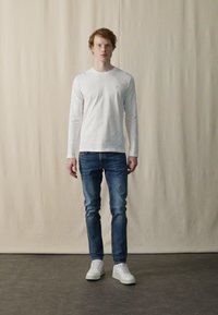 Hvid langærmet skjorte, blå jeans og hvide sneakers. Modellen står foran en neutral baggrund og fremviser det enkle, afslappede outfit.