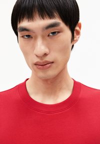 Junge Person mit kurzem schwarzen Haar, die ein rotes Rundhals-Shirt trägt, neutrale Miene, einfarbig weißer Hintergrund.