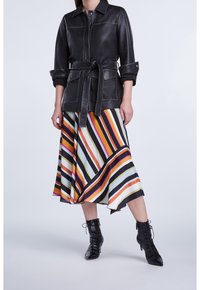 Veste ceinturée en cuir noir avec fermeture à boutons, portée sur une jupe midi rayée multicolore présentant des motifs diagonaux et des bottes noires à lacets.