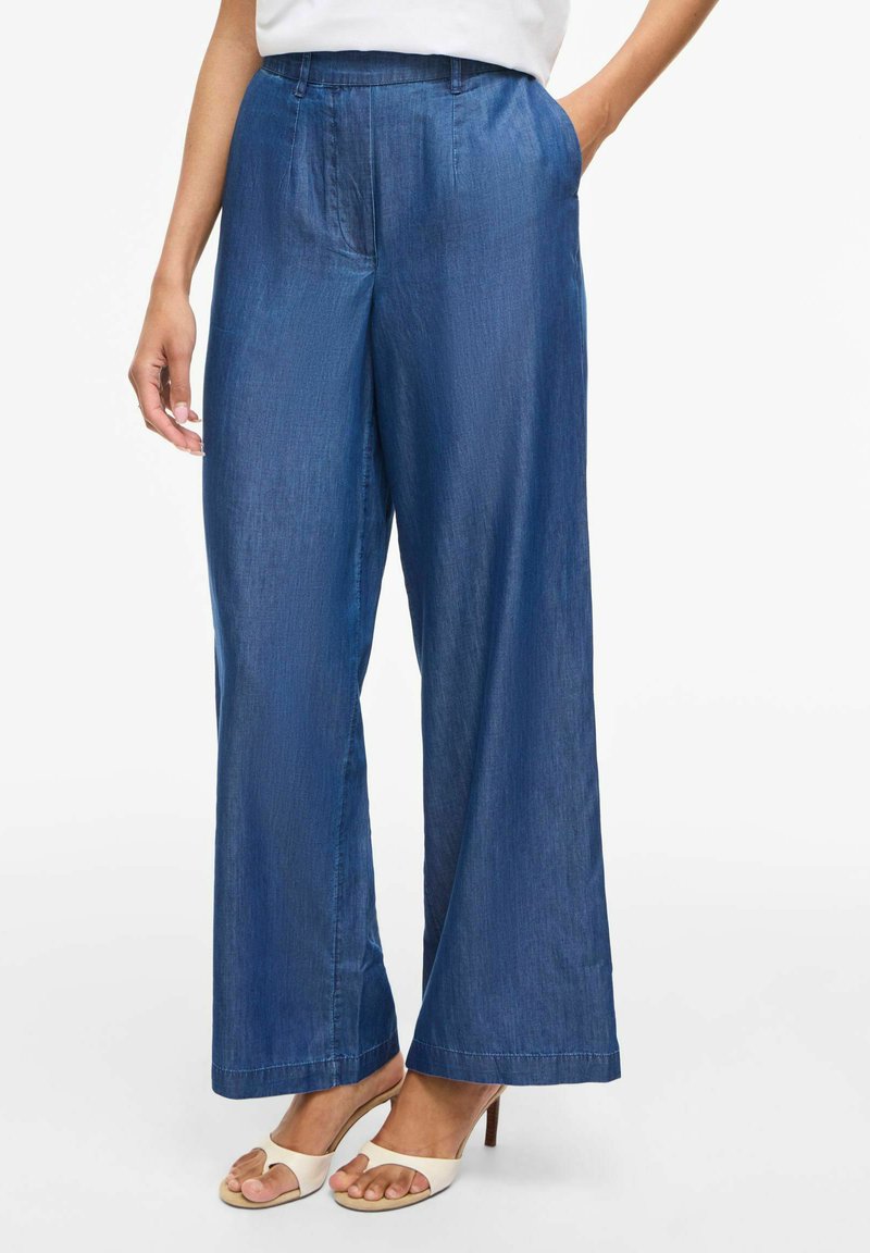 Pantaloni largi din denim albastru purtați cu sandale albe cu vârf deschis, mâna dreaptă a modelului în buzunar, fotografiați de la talie până la picioare.