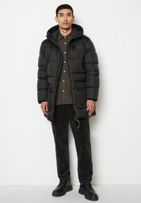 Giacca a vento nera con cappuccio, caratterizzata da un design trapuntato, bottoni frontali e chiusura con zip. Indossata sopra una camicia a scacchi e pantaloni scuri.