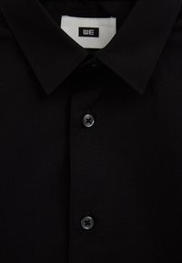 Melna pogājama krekla ar apkakli, vienkārša dizaina, ar divām melnām pogām un zīmola etiķeti apkakles iekšpusē.