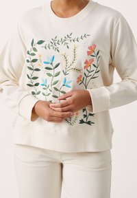 Sweatshirt beige avec une broderie florale colorée en bleu, vert, orange et jaune ; texture douce ; coupe décontractée avec des poignets et un ourlet côtelés.
