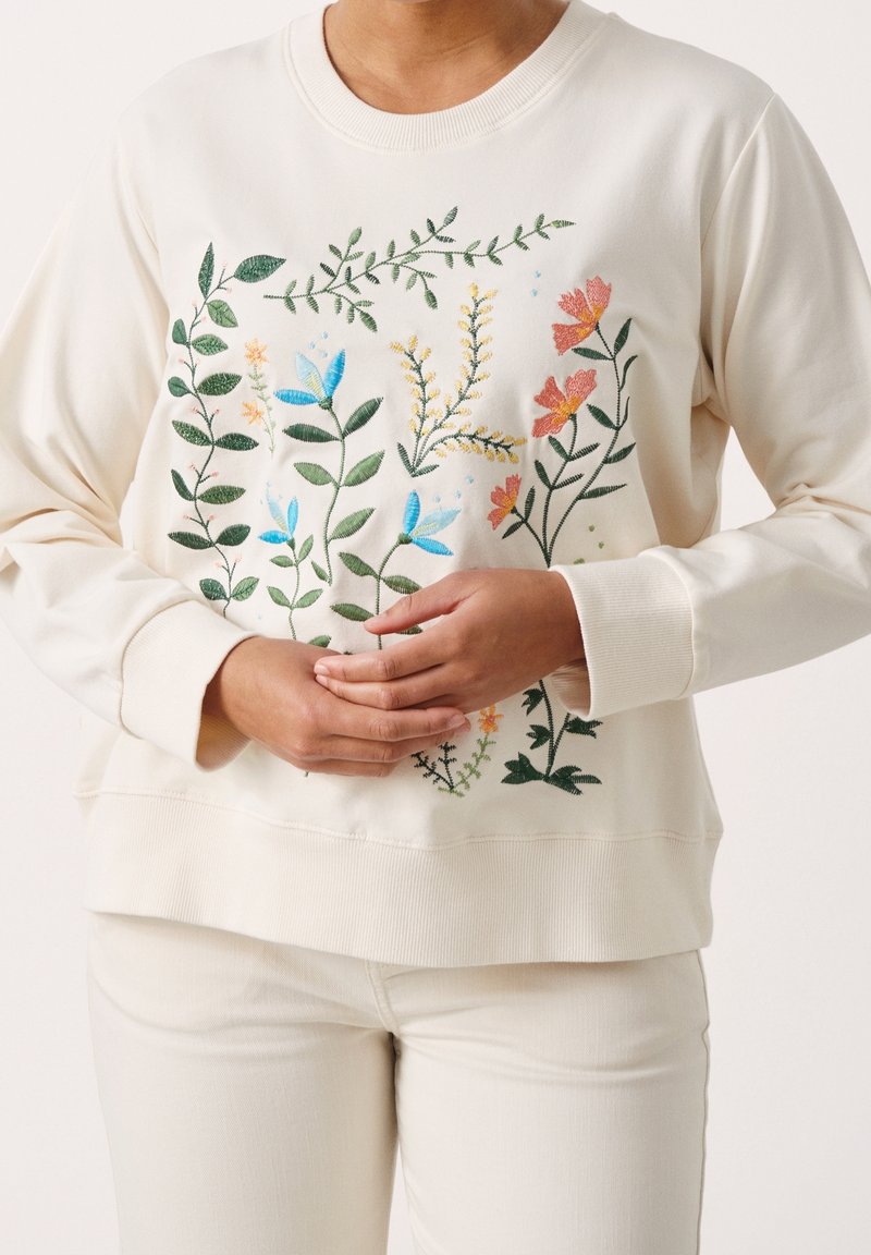 Sweatshirt beige avec une broderie florale colorée en bleu, vert, orange et jaune ; texture douce ; coupe décontractée avec des poignets et un ourlet côtelés.