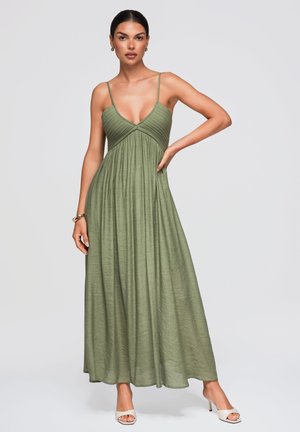 Femme portant une robe longue sans manches vert olive avec de fines bretelles et des sandales blanches à talons, debout avec une main sur la hanche.