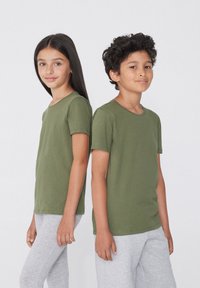 Děti stojí zády k sobě, oblečeni ve svetrech s krátkým rukávem olivově zelené barvy a šedých legínách. Materiál vypadá hladce a přiléhavě.