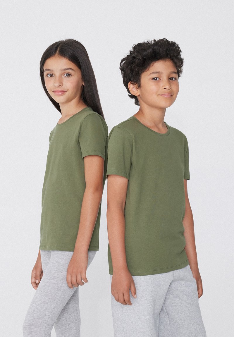 Děti stojí zády k sobě, oblečeni ve svetrech s krátkým rukávem olivově zelené barvy a šedých legínách. Materiál vypadá hladce a přiléhavě.