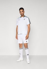 Mand iført hvid Adidas fodbolduniform med stribet trøje, stribede sokker og fodboldstøvler, stående på en enkel lys grå baggrund.