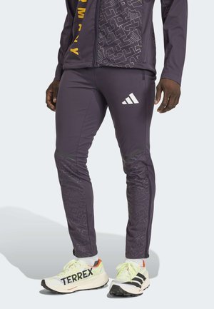 Leg wearing purple athletic pants with Adidas logo and white Terrex running shoes with black stripes and orange heel accents.