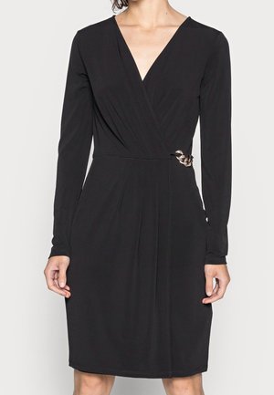 Femme portant une robe portefeuille noire à manches longues avec un profond décolleté en V et un détail chaîne en or à la taille.