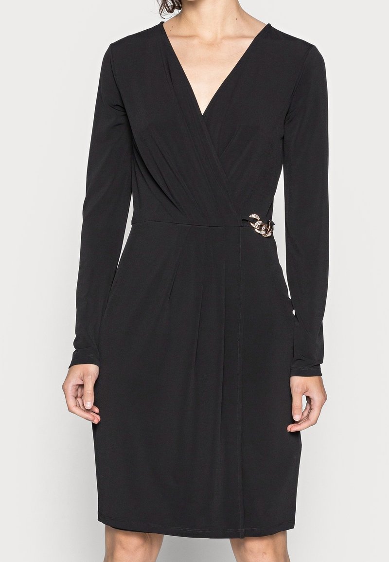 Femme portant une robe portefeuille noire à manches longues avec un profond décolleté en V et un détail chaîne en or à la taille.
