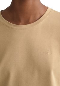Beige t-shirt med kort ärm, tillverkad av mjukt tyg, med rundad halsringning och präglad logotypdetalj på bröstet.