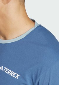 Maglietta sportiva blu con collo azzurro chiaro, realizzata in tessuto morbido. Presenta il logo bianco "TERREX" sul davanti, trama liscia e maniche corte.