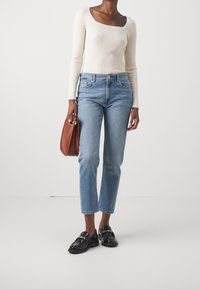 Licht beige geribbelde lange mouwen top, blauwe rechte jeans, zwarte instapschoenen met gouden hardware en een bruine tas. Eenvoudige, moderne outfit.