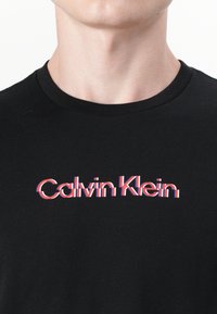 Black cotton t-shirt with a round neck. Features a distorted "Calvin Klein" logo in red, white, and blue lettering across the chest.