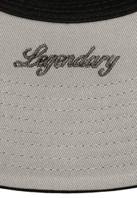 Tejido gris con texto negro bordado que dice "Legendario" encima de filas de detalles cosidos, parte del ala de una gorra de béisbol con ribete negro.