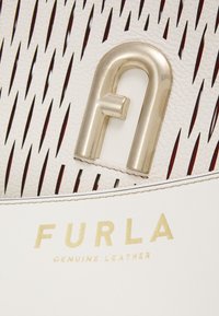 Bílá kožená taška s vystřiženým designem, zlatými detaily a embosovaným logem "FURLA." Má hladkou texturu a kontrastní barvu vnitřní části.