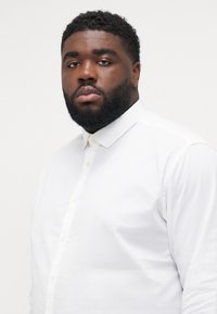 Chemise blanche à manches longues avec un col, présentant une texture lisse et des boutons sur le devant.