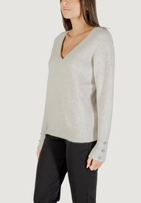 Pullover grigio chiaro a scollo a V con texture a coste e dettagli metallici. Presenta una vestibilità ampia e bottoni sulle maniche. Abbinato a pantaloni neri.