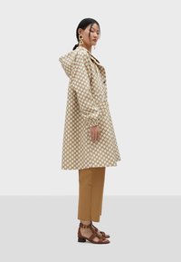 Beżowy płaszcz przeciwdeszczowy w kropki, z kapturem i zapinany na guziki, w połączeniu z brązowymi spodniami typu cropped i brązowymi sandałami na obcasie.