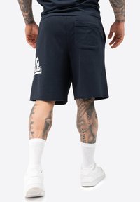 Mørkeblå bomulds shorts med en afslappet pasform, der har en baglomme og en kantet kant. Sammen med hvide sneakers og sokker.