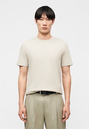 Fiatal férfi fekete rövid hajjal, világos bézs rövid ujjú pólót visel, amelyet bézs, redős nadrágba tűrt be, fekete övvel.