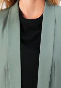 Krk a horní část hrudníku osoby v černé košili a světle zeleném saku, volné vlasy viditelné po obou stranách.