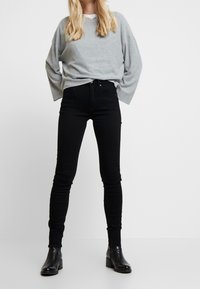 Sudadera gris claro de gran tamaño con escote en V. Vaqueros ajustados negros con textura suave. Botines negros con tacón bajo y grueso.