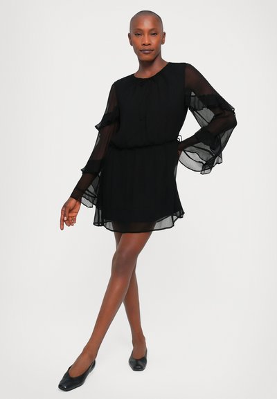 Black chiffon dress with long, sheer ruffled sleeves, gathered waist, and a flowing skirt. Paired with black flats. Smooth, lightweight fabric.