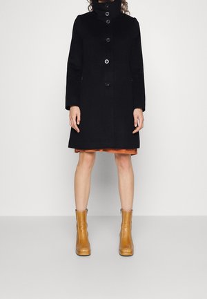 Manteau en laine noir avec un col haut, comportant six boutons et des manches longues. Porté par-dessus une robe couleur rouille et associé à des bottines beiges.