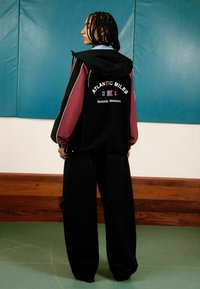 Haut à capuche noir et bordeaux à blocs de couleur avec le texte blanc "ATLANTIC MILES" et "Seaside Mariners," associé à un pantalon large noir.