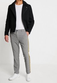 Černá přiléhavá bunda s mírně zvýšeným límcem; doplněná šedými károvanými kalhotami se žlutým postranním pruhem; bílé tenisky.