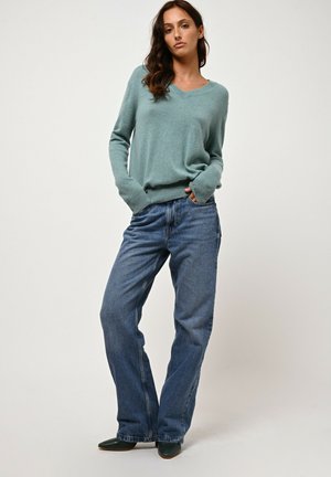Maglione a V leggero color teal, texture morbida, abbinato a jeans blu scuro a vestibilità ampia, tasche visibili, indossato con scarpe verdi scure.