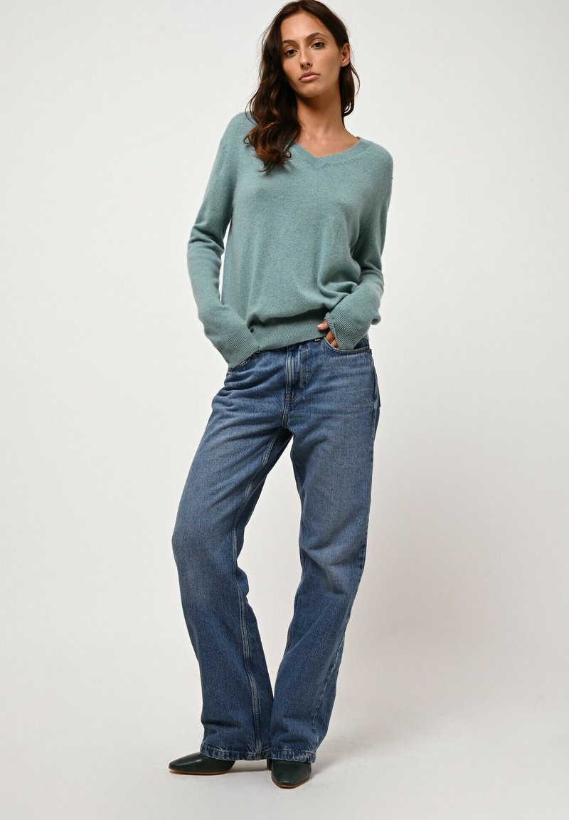 Hellblauer V-Ausschnitt-Pullover mit weicher Textur, kombiniert mit lockeren, dunkelblauen Jeans, sichtbare Taschen, getragen mit dunkelgrünen Schuhen.