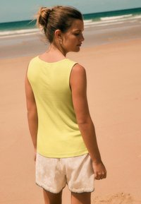 Débardeur jaune à côtes associé à un short blanc avec un motif de feuilles de palmier et des finitions noires, sur fond de plage de sable.