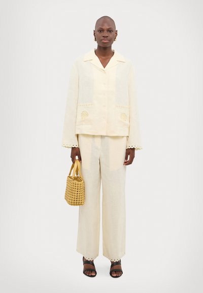 Ensemble en lin à deux pièces de couleur jaune pâle ; veste courte à boutons avec poches et accents brodés, associée à un pantalon large. Sac tissé jaune.