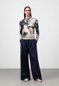 Lange mouw top met een blauw en crème abstract patroon, gecombineerd met losse donkere denim broek en crème instapschoenen.