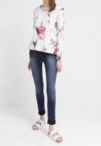Kjortärmad blus med blommönster i vitt med rosa och gröna accenter, matchat med mörkblå skinny jeans och vita sandaler med spännen.
