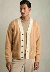 Cardigan beige clair avec des rayures blanches, doté d'un grand col blanc, de boutons et de poches avant. Porté par-dessus un pull beige.