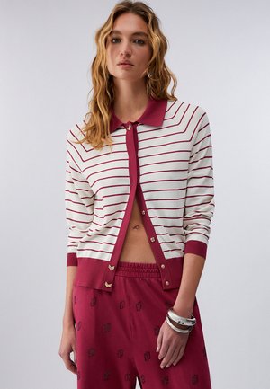 Giovane donna con un cardigan bianco a righe bordeaux, parzialmente allacciato, pantaloni bordeaux con motivi e braccialetti d'argento.