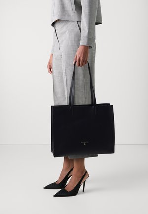 Person in kariertem Anzug mit großer schwarzer Tragetasche mit dünnen Griffen, trägt schwarze spitze High Heels, steht vor weißem Hintergrund.