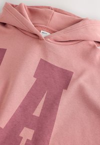 Sweat à capuche rose avec une grande lettre "A" rose foncé imprimée sur le tissu et une étiquette visible indiquant "NEXT" à l'intérieur de la capuche.