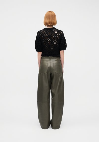 Γυναίκα με κοντά κόκκινα μαλλιά που φοράει ένα μαύρο πλεκτό τοπ με κοντά μανίκια και ψηλόμεσα φαρδιά παντελόνια σε ελιά πράσινο, όρθια με την πλάτη προς την κάμερα.