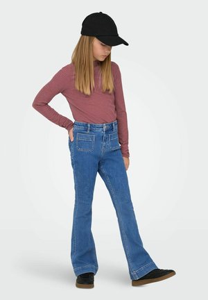 Lány fekete sapkában, rózsaszín hosszú ujjú pólóban, elöl zsebes kék farmerral és fekete cipőben, egy kezét a zsebében tartva, lefelé nézve áll.