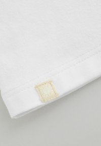 Witte katoenen T-shirt hoek met een textuur van een tan kleurige label genaaid op de zoom, met een ronde vorm en subtiele stiksels.