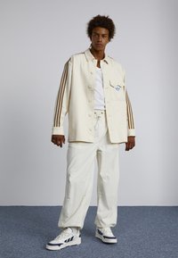 Ung man står klädd i en krämfärgad jacka med bruna ränder på ärmarna, vit T-shirt, lösa vita byxor och vita sneakers med marinblå detaljer.