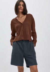Cardigan en tricot marron avec un col en V et une fermeture à boutons, associé à un short gris foncé avec une taille élastique et des poches.