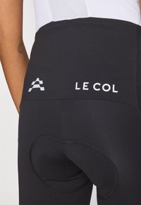 Melni riteņbraukšanas šorti ar gludu tekstūru, kuros ir izteikts balts "LE COL" logo un;apaļa spilvena detaļa aizmugurē.