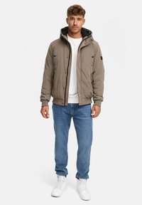 Brun yderjakke med hætte, lynlåslukning foran og sort indvendig foring, parret med blå jeans og hvide sneakers.
